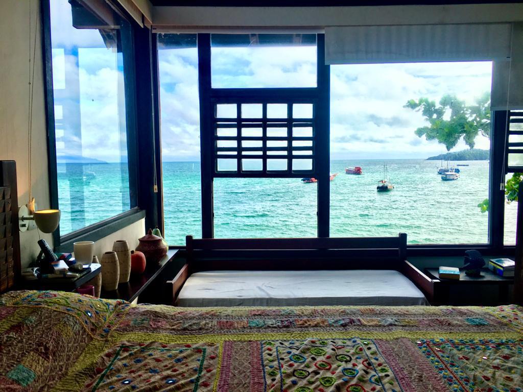 A bedroom is highlighted by a large window overlooking the ocean, with boats anchored in the distance. A bed with a colorful patterned blanket is positioned near the window, allowing natural light to fill the space. Decorative items are arranged on nightstands.