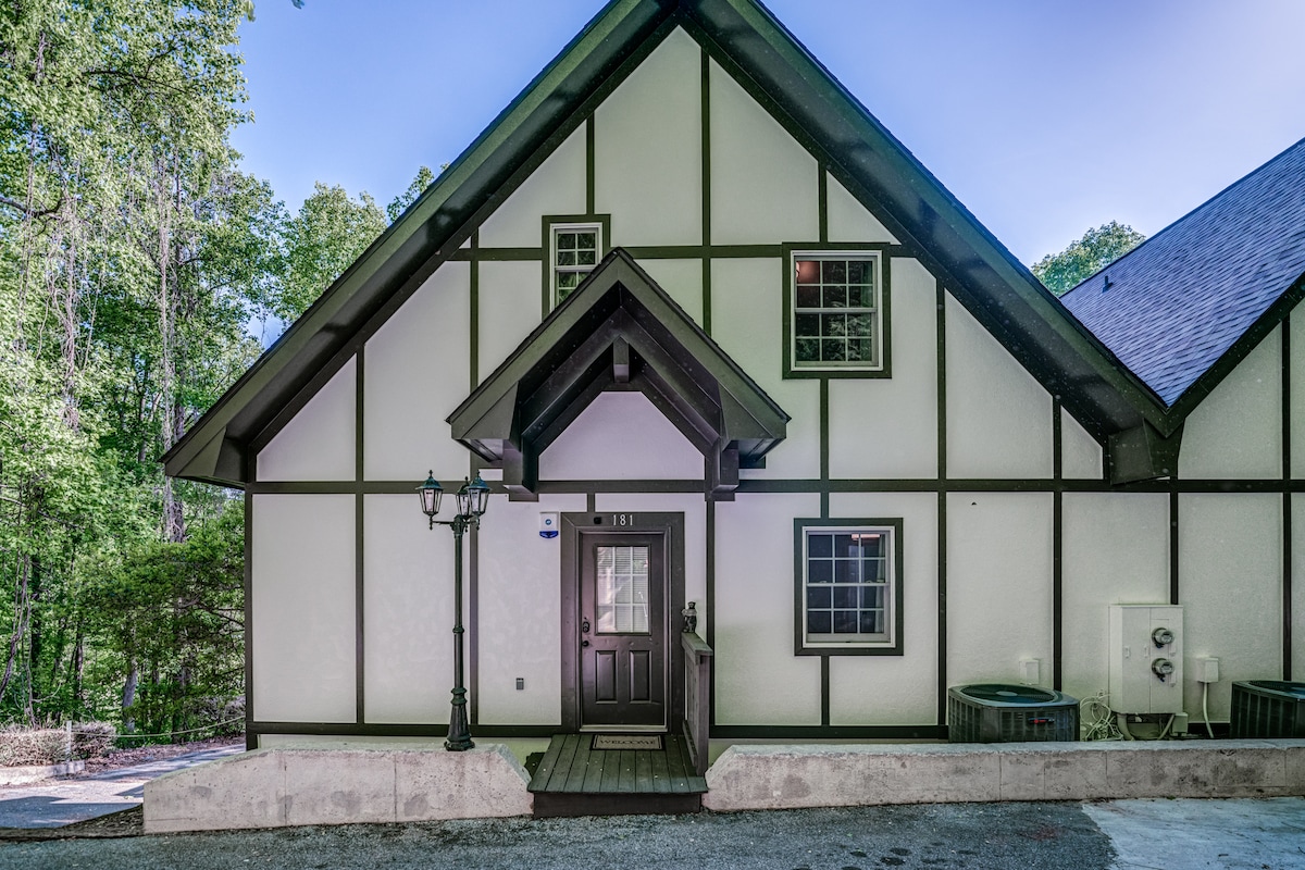 The exterior of the townhouse features a charming A-frame design with a combination of white stucco and dark wood accents. A welcoming entrance is framed by a gabled roof, with large windows allowing natural light. The space includes outdoor elements such as a hot tub and split-level access.