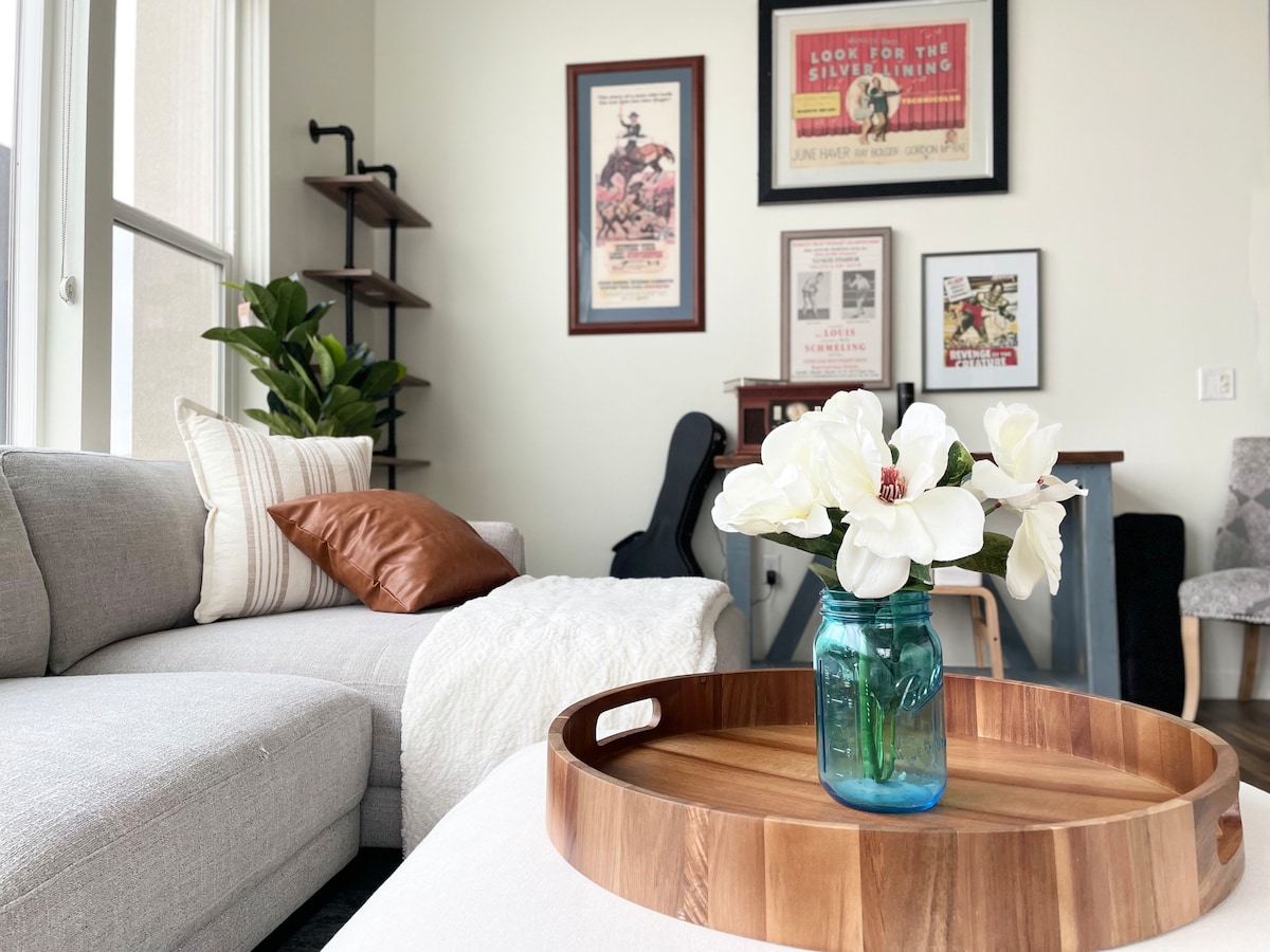 A cozy living space is featured, showcasing a light grey sectional sofa adorned with throw pillows. A wooden tray holds a blue vase containing white flowers. Art prints hang on the wall, complemented by a small indoor plant nearby.