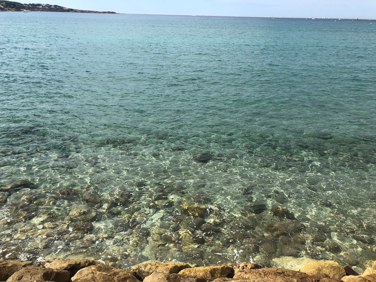 Clear waters reveal a rocky seafloor, with gentle waves lapping against the shore. The tranquil blue hues of the sea extend to the horizon, where the sky meets the water. Sunlight glistens on the surface, creating a serene and inviting atmosphere.