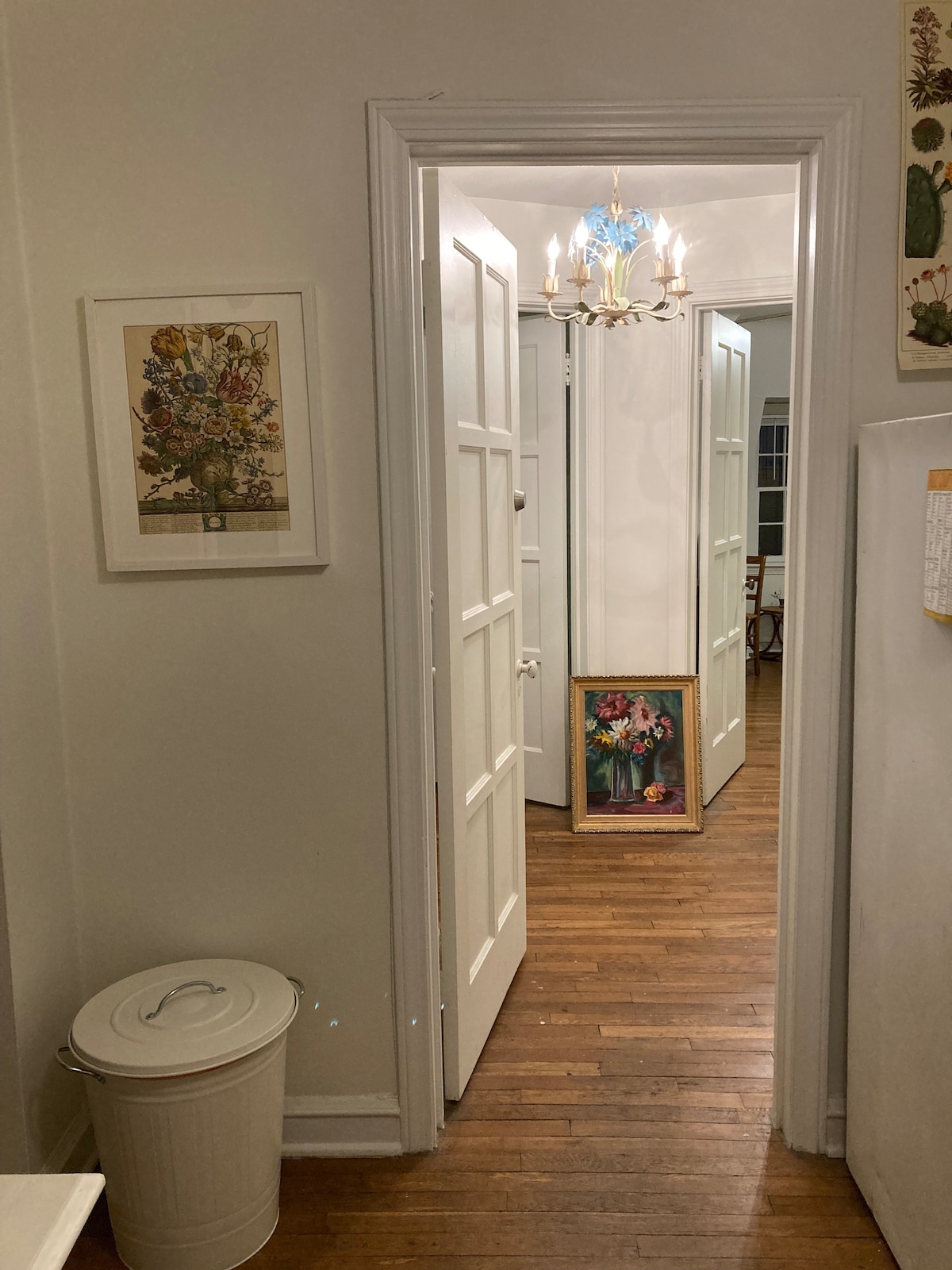 A view of a hallway features bright white walls and wooden flooring. A small waste bin is positioned to the left. An ornate chandelier hangs from the ceiling, illuminating the space. Colorful artwork is displayed at the end of the hallway, adding a touch of charm.
