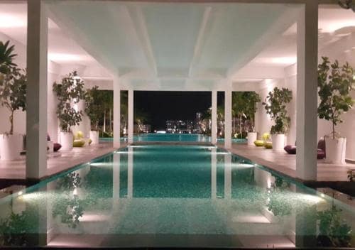 An elegant outdoor swimming pool is illuminated by soft lighting, reflecting a serene atmosphere. Lush greenery is strategically placed in white planters along the edges, with contemporary lounge seating providing space for relaxation. The view of the city skyline enhances the tranquil setting.