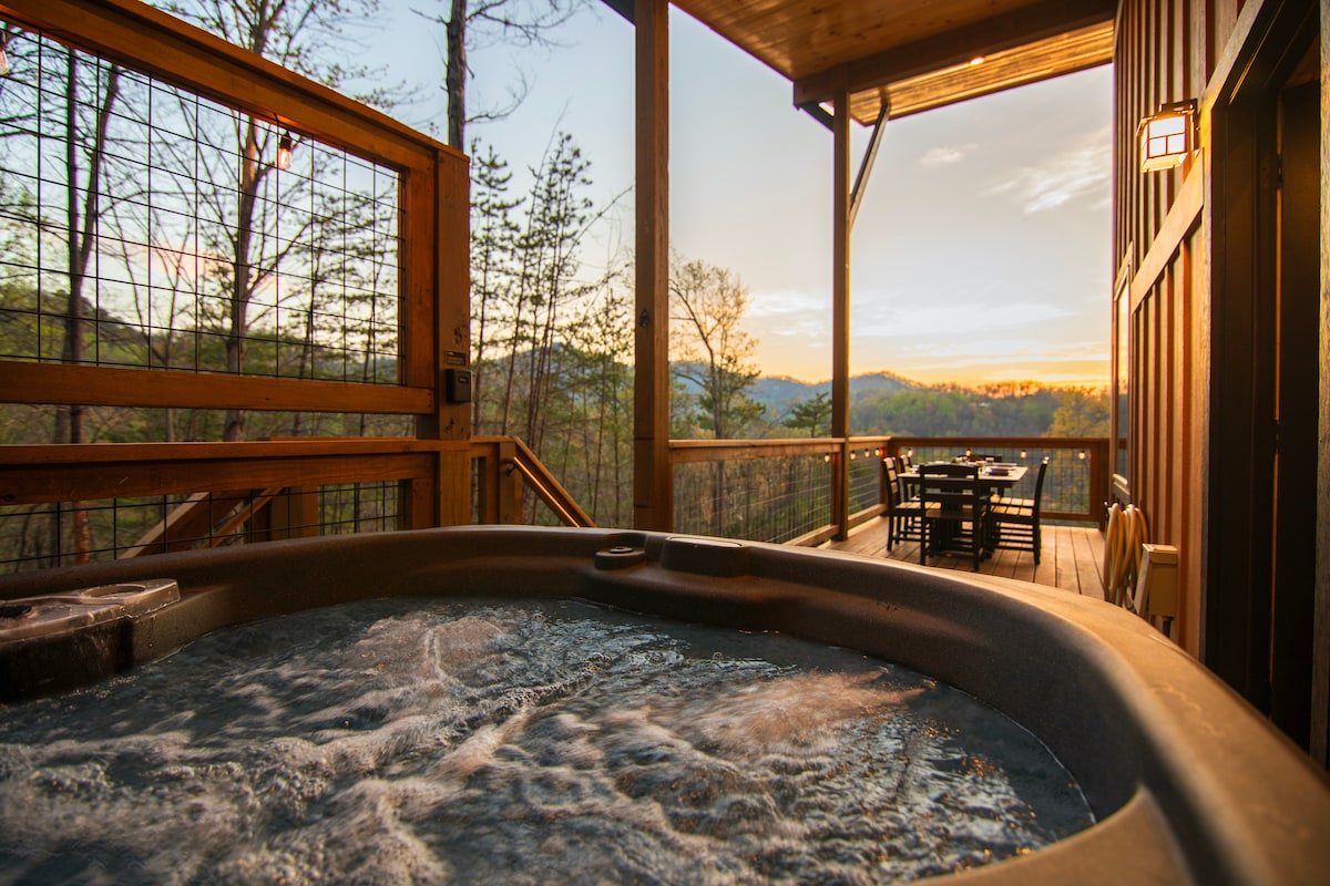 A private hot tub is positioned on a covered deck, with steam rising gently from the water. The surrounding natural landscape shows trees and rolling mountains, while a dining area with table and chairs is visible in the background, illuminated by a warm sunset.