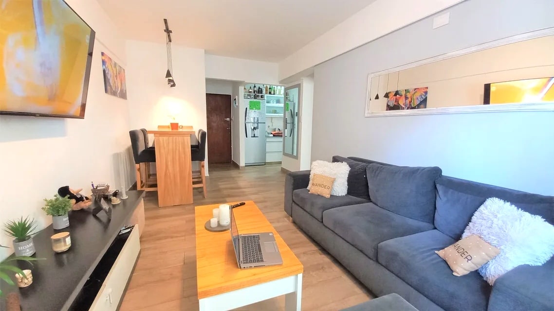 A modern living area features a comfortable grey sofa with decorative pillows. A wooden coffee table is centrally placed, accompanied by a small plant and a laptop. A wall-mounted TV and a dining area are visible, showcasing an open layout and inviting ambiance.