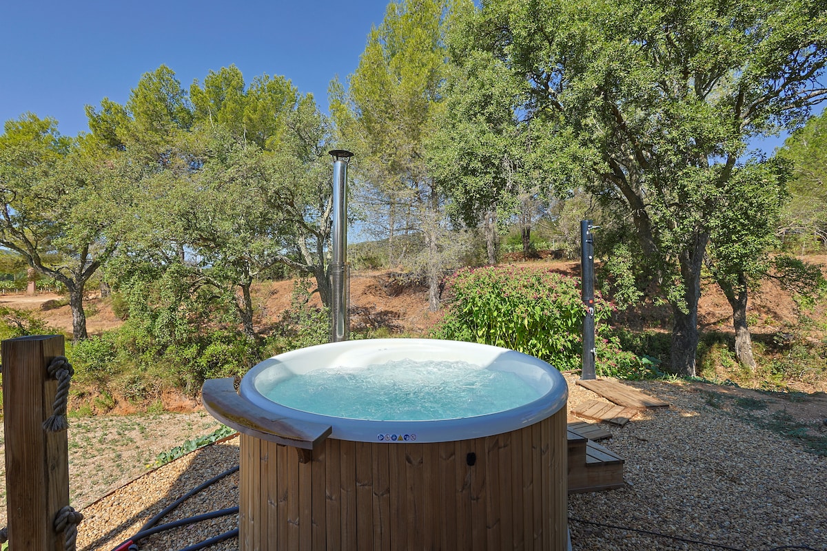 A wooden hot tub is situated outdoors, surrounded by lush greenery and trees. Natural light highlights the clear water, inviting visitors to relax. A stone path leads to the tub, blending harmoniously with the landscape.