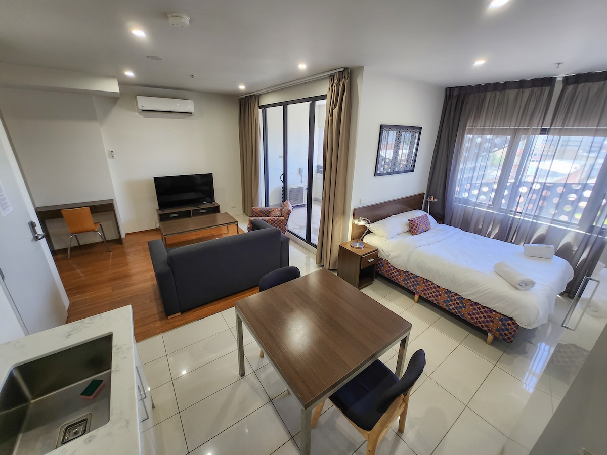 A modern studio layout is visible, featuring a spacious bed accompanied by a bedside table. A cozy seating area with a loveseat and armchair is present, while a dining table for four sits nearby. Large windows provide ample natural light and access to an outdoor area.