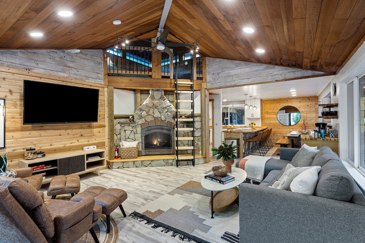 A spacious living area features a cozy seating arrangement, including a large sectional sofa and armchairs. A stone fireplace acts as a focal point, while large windows invite natural light. A ladder leads to an upper loft, contributing to the cabin's open layout and warmth.