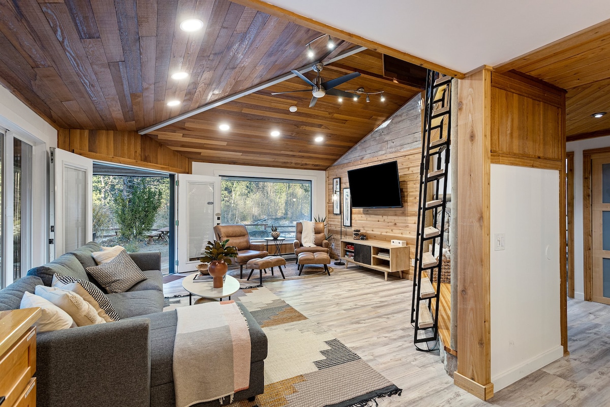 A spacious living area is highlighted by wooden ceilings and floors, complemented by modern furnishings. A large sectional sofa is positioned around a coffee table, with two armchairs nearby. A flat-screen TV is mounted on the wall, and large windows offer views of the surrounding nature.