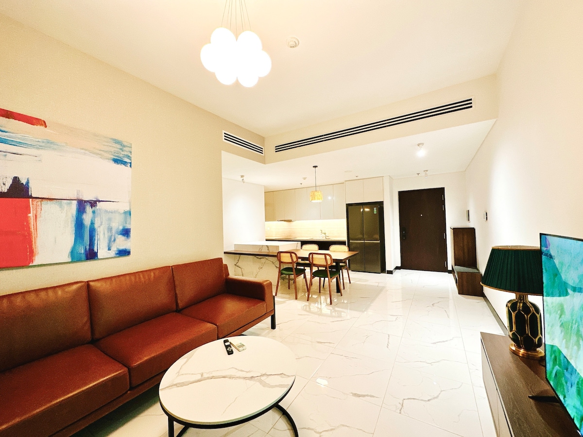 The living area features a modern brown leather sofa arranged around a circular coffee table. A vibrant piece of wall art adds color to the neutral tones. A dining space with wooden chairs is visible, along with a well-equipped kitchen area in the background.