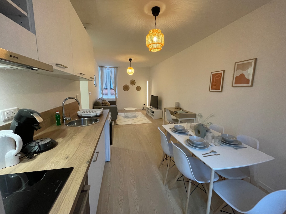 An open kitchen and dining area features a long white table set for six with stylish dishware. The kitchen has modern appliances and wooden countertops. A cozy living space is visible in the background, complete with a soft rug and tasteful wall art.