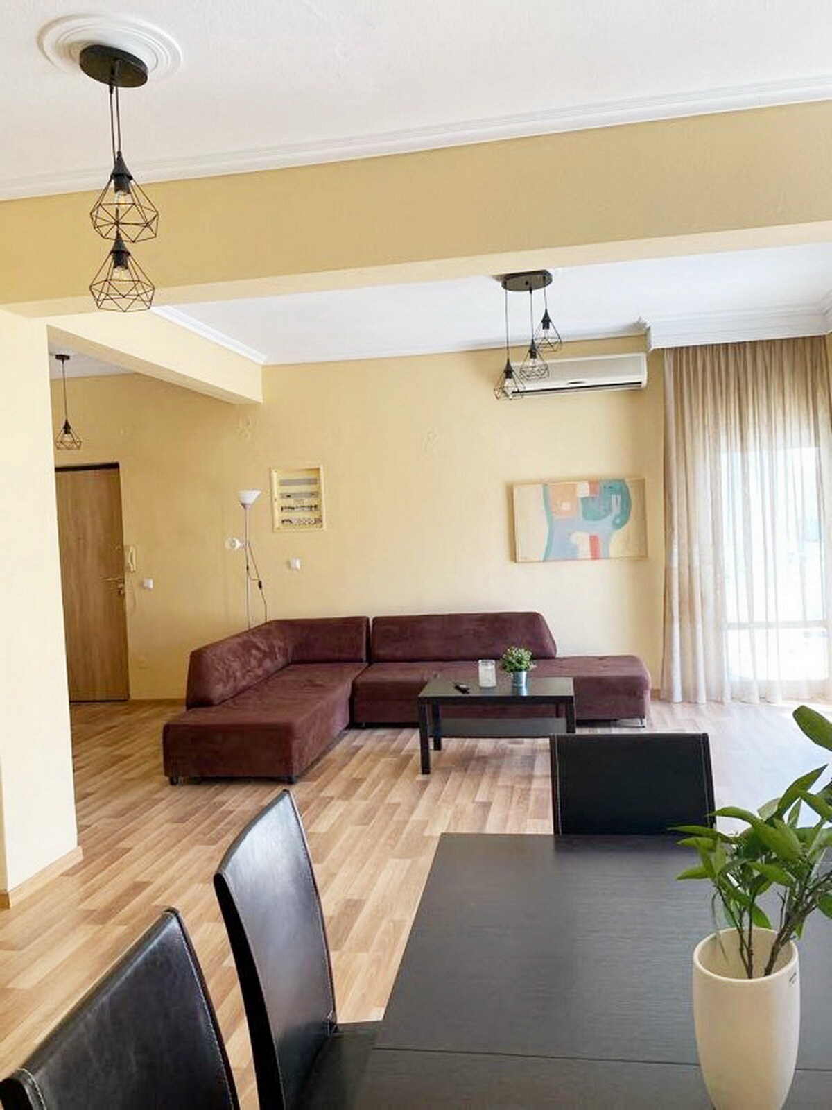 A spacious living area features a brown sectional sofa, a coffee table, and a light-colored wooden floor. Large windows with sheer curtains allow natural light to fill the space, while a decorative plant and artwork on the walls add subtle accents.