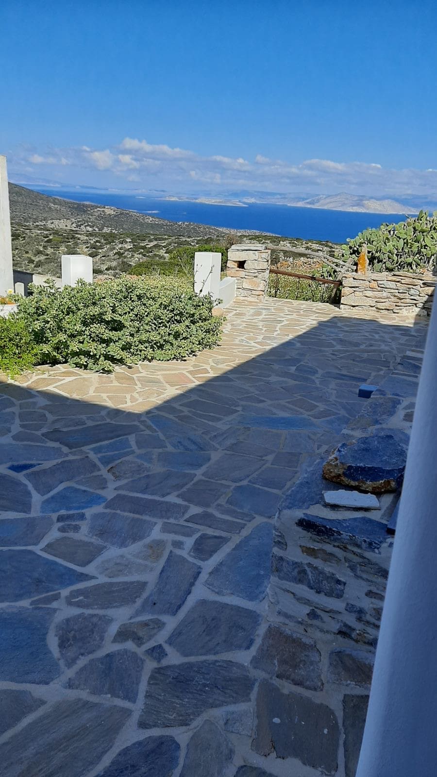 A stone pathway leads through a landscaped area, bordered by low greenery and featuring natural stone textures. The expansive view of the sea is visible in the distance, accompanied by a clear blue sky. Sunlight creates contrasting shadows on the pathway.