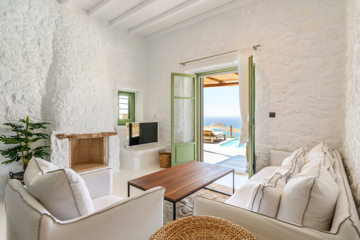 A bright and airy living area features white stone walls and ceiling beams. A comfortable seating arrangement is set around a wooden coffee table, with greenery positioned nearby. Open doors lead to an outdoor terrace with views of the sea, inviting natural light into the space.