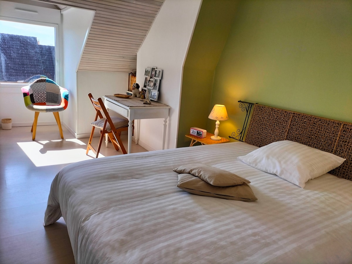 A light-filled bedroom features a spacious bed with soft white linens and pillows. A decorative headboard adds character, while a small desk and chair provide a workspace. A bedside lamp and wall art enhance the cozy atmosphere, creating an inviting space.