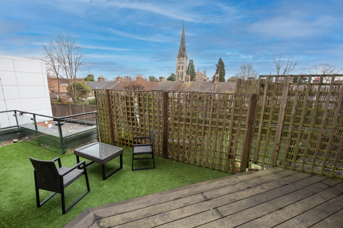 An outside space features two chairs and a glass-top table on a wooden deck surrounded by artificial grass. A wooden lattice provides privacy, and a nearby church steeple is visible against the blue sky, enhancing the neighborhood ambiance.