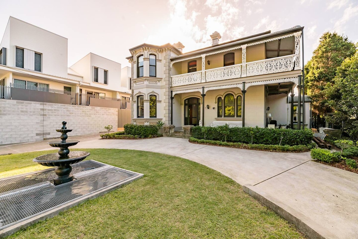 An elegant mansion is showcased, framed by a manicured lawn and paths. A decorative fountain is centered in the foreground. Architectural details, such as a balcony and ornate features, highlight the home's historic charm, while modern structures are visible in the background.
