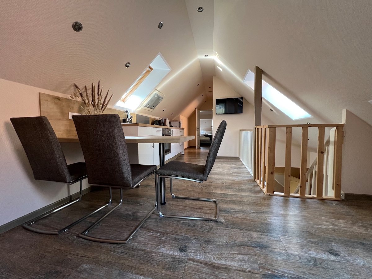The dining area is furnished with a modern table and two sleek chairs, situated in a bright, open space. Natural light filters through skylights and adds warmth to the wood-tone flooring. A staircase and kitchen area are visible in the background.