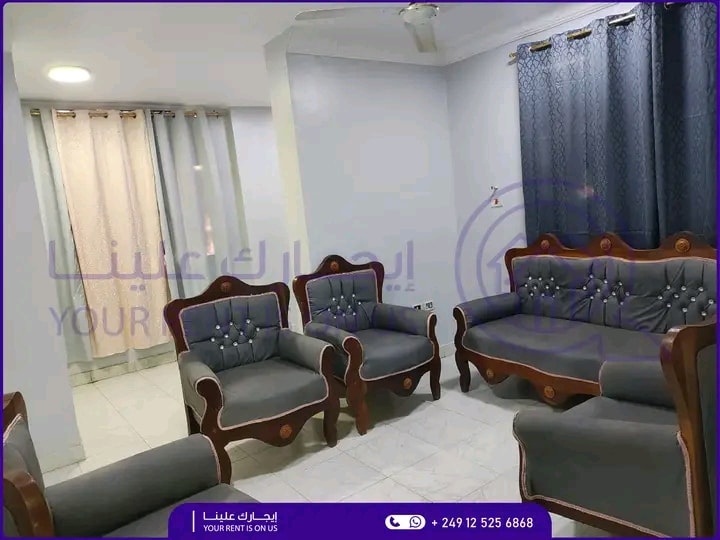 A comfortable seating area is furnished with an arrangement of soft, grey upholstered chairs and a settee, each featuring decorative button detailing. Light curtains frame a window, allowing gentle light to brighten the space.