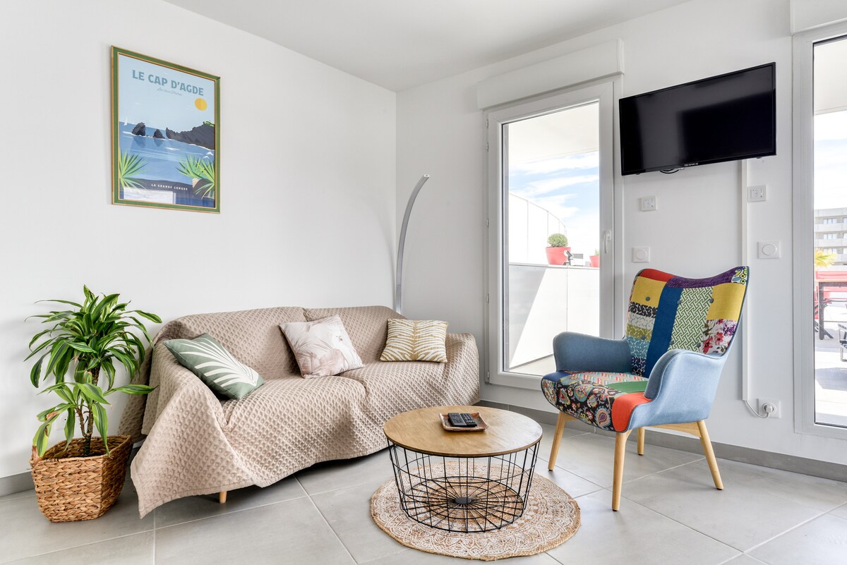 A bright living area is featured, with a beige couch adorned with soft pillows and a colorful armchair. A round coffee table sits on a neutral area rug, and large windows allow natural light to enhance the space. A wall-mounted TV is positioned for viewing.