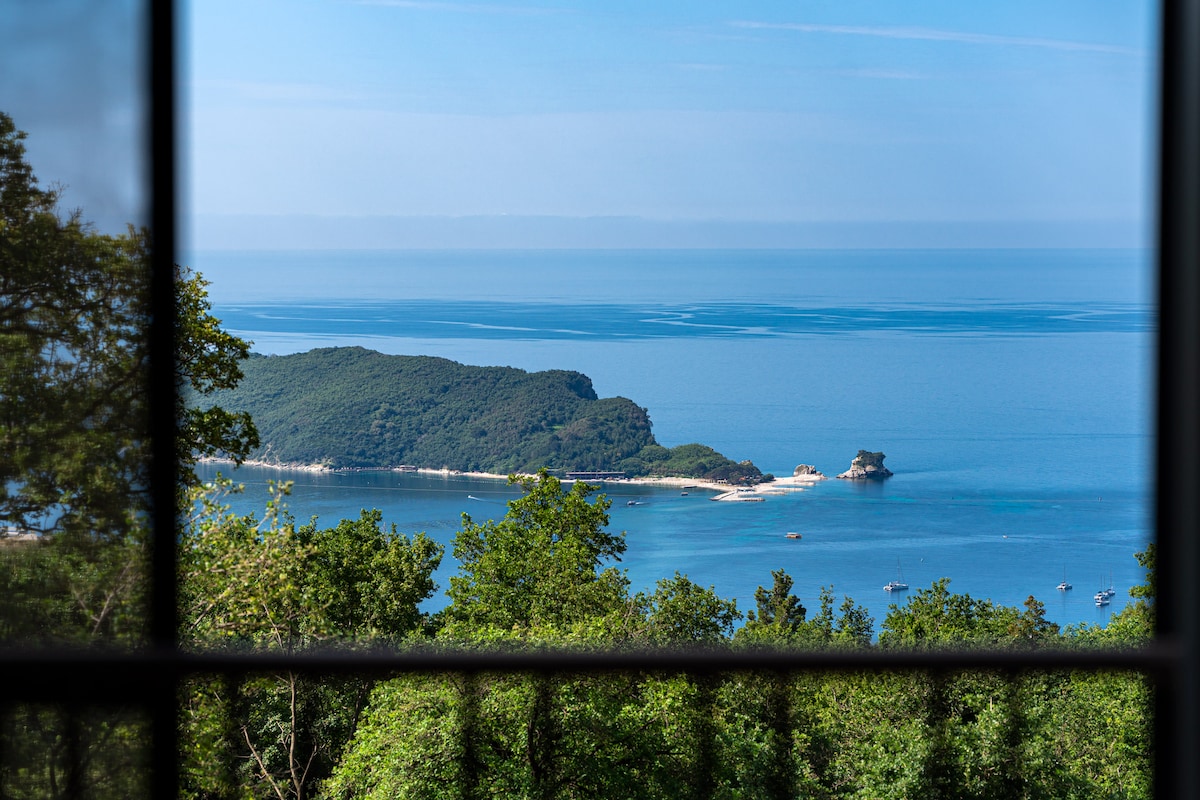 A panoramic view of the Adriatic Sea is framed by a window, showcasing a lush green landscape that borders the serene water. Gentle waves can be seen in the distance, alongside small boats dotting the coastline, contributing to a peaceful ambiance.