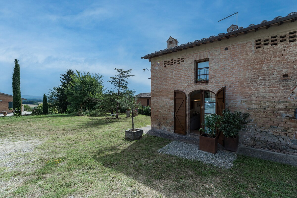 The exterior of the rustic two-story building features large wooden doors flanked by potted plants. The surrounding grassy area is punctuated by trees and provides open space. A backdrop of the serene countryside is visible under a clear blue sky.
