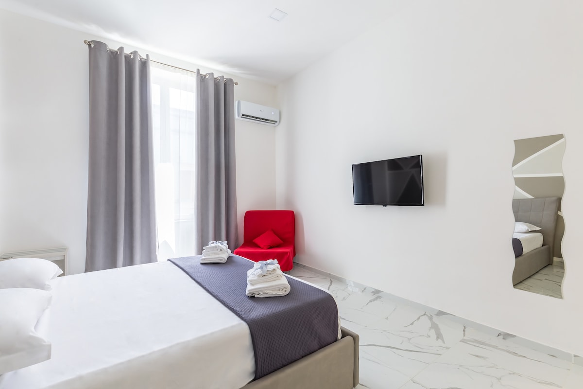 A well-lit bedroom features a spacious bed with neatly arranged towels. A contemporary chair in red is placed near a large window, which is dressed with light gray curtains. A flat-screen television is mounted on the wall, complementing the room's minimalist design.