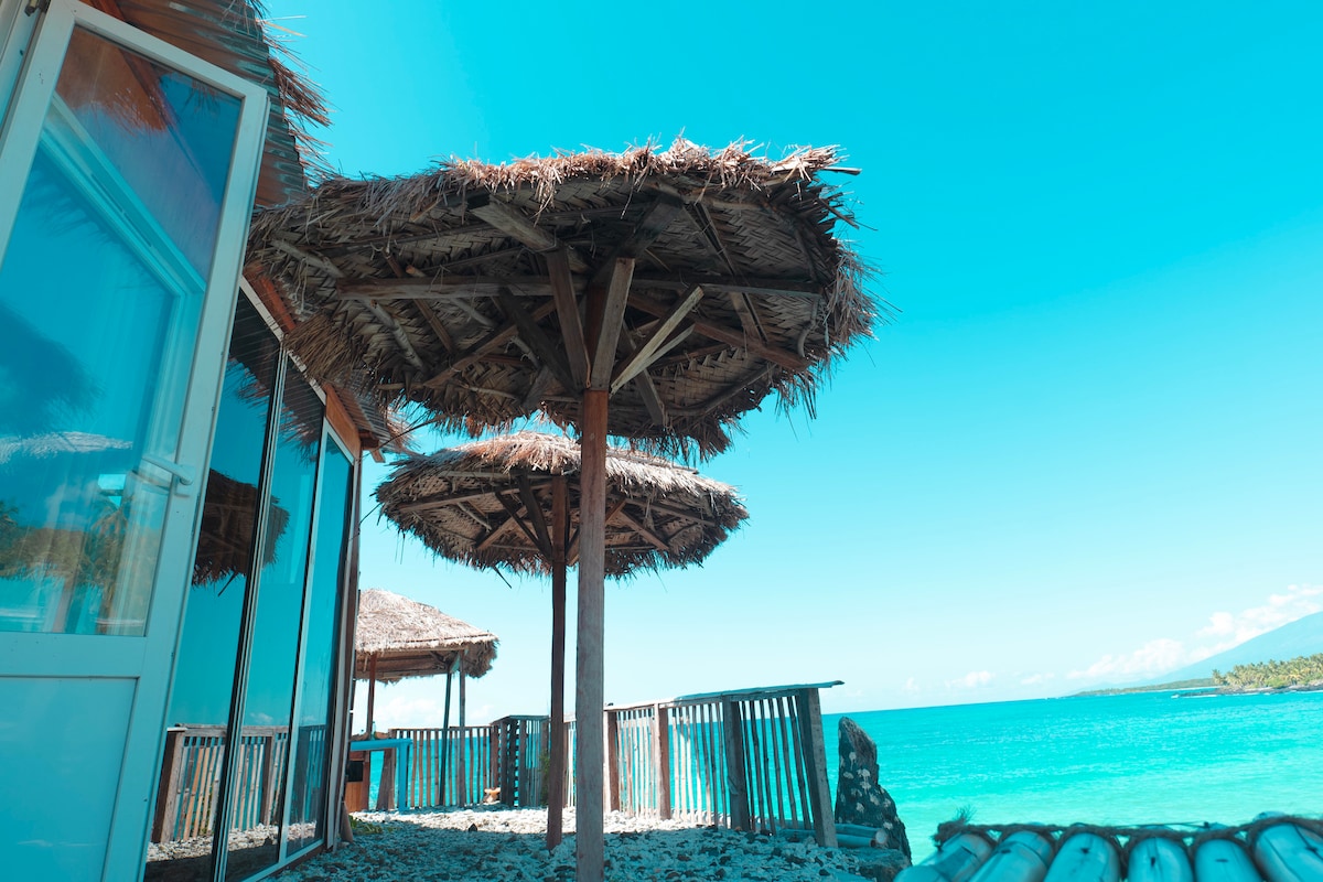 Two thatched umbrellas provide shade on a sandy area near the bungalow. The clear blue sea and sky create a vibrant backdrop, while the structure's glass panels reflect natural light, enhancing the serene coastal setting.
