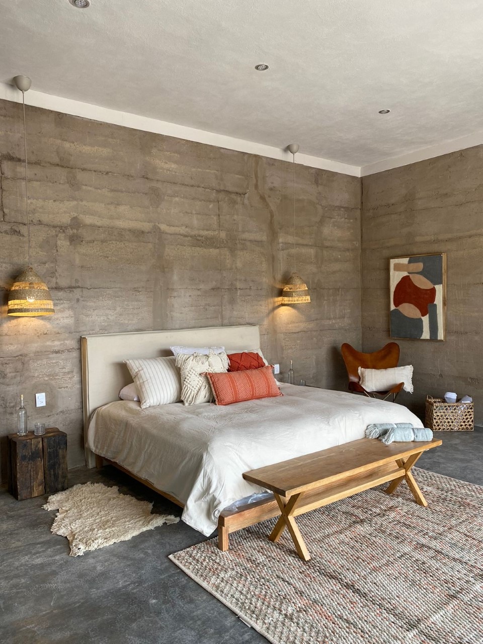 A spacious bedroom features textured rammed earth walls and soft lighting from two hanging lamps. A large bed is dressed in neutral linens with accent pillows in warm tones. A wooden bench adds functional seating in front of the bed, and a woven rug enhances the earthy aesthetic.