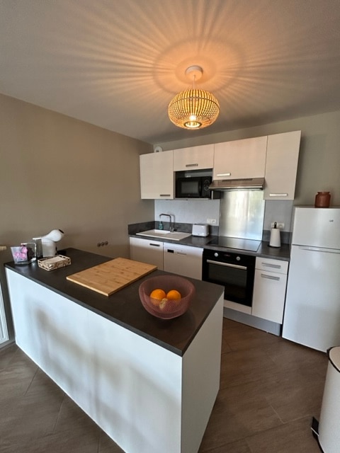 The kitchen features a modern design with sleek cabinetry and a central island. A pendant light creates a warm glow, illuminating the countertop. Equipped appliances, including a refrigerator and oven, are visible, alongside a bowl of oranges adding a touch of color.