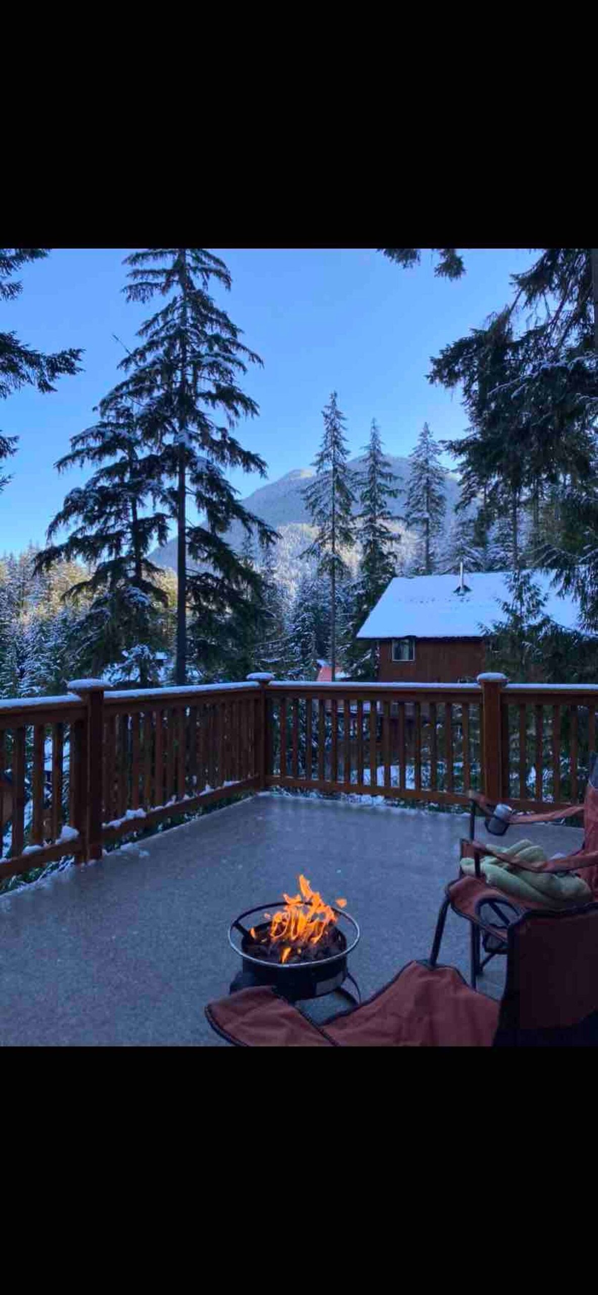 A spacious deck is shown, featuring a propane fire pit with a flickering flame. Snow-covered trees surround the cabin, while distant mountains rise under a clear blue sky, providing a serene winter landscape.