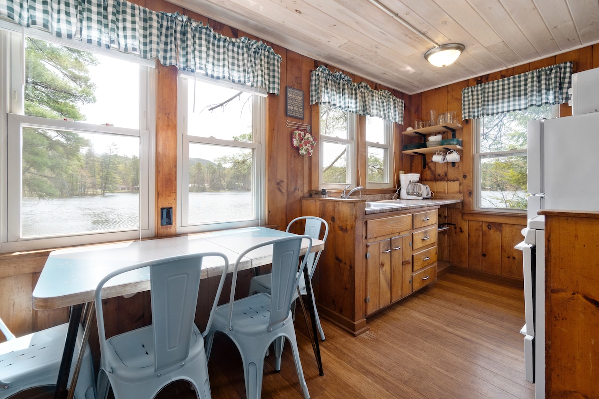 A quaint eat-in kitchen is illuminated by natural light filtering through multiple windows, offering views of Lake Vanare. Wooden paneling and checkered curtains complement the space. A table with blue chairs and wooden cabinetry enhance the inviting atmosphere of this functional area.