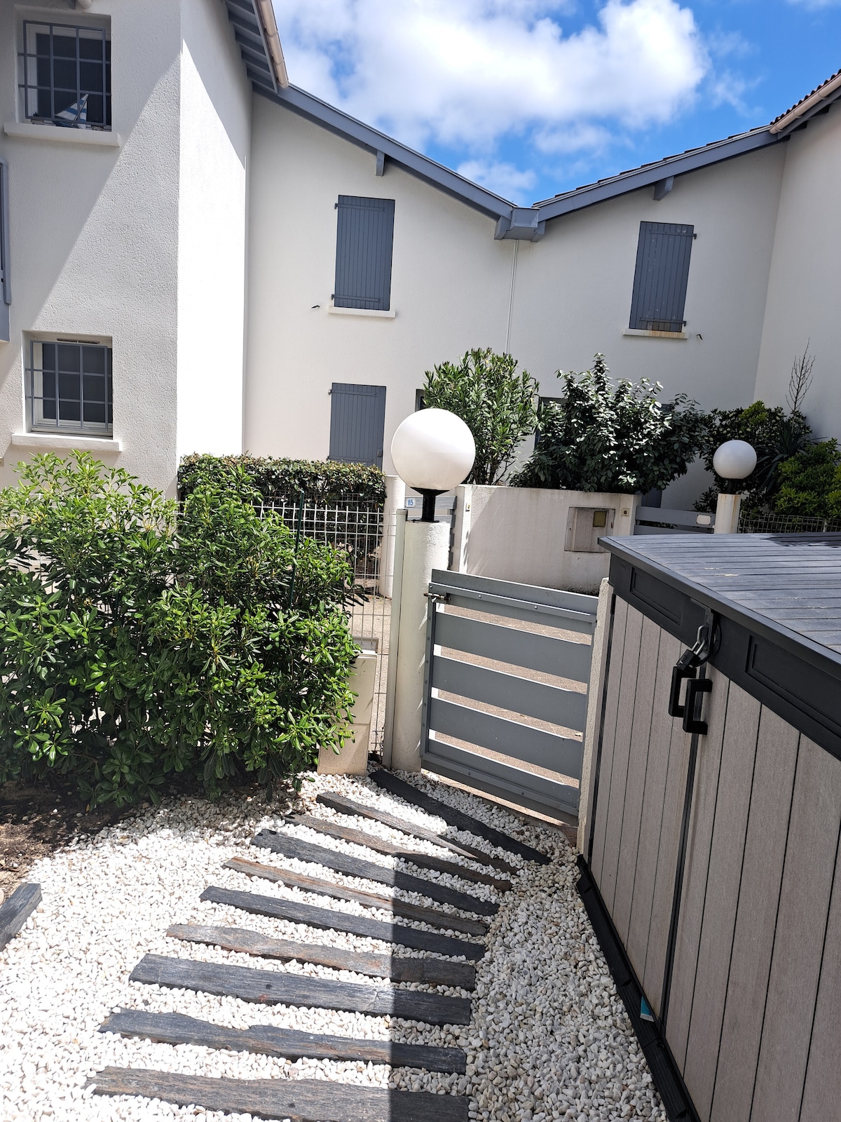 A well-maintained pathway with stone steps leads to a gate, surrounded by lush greenery and neatly arranged gravel. The light fixtures along the pathway enhance visibility. The white-walled buildings in the background feature windows with shutters, creating a uniform aesthetic.