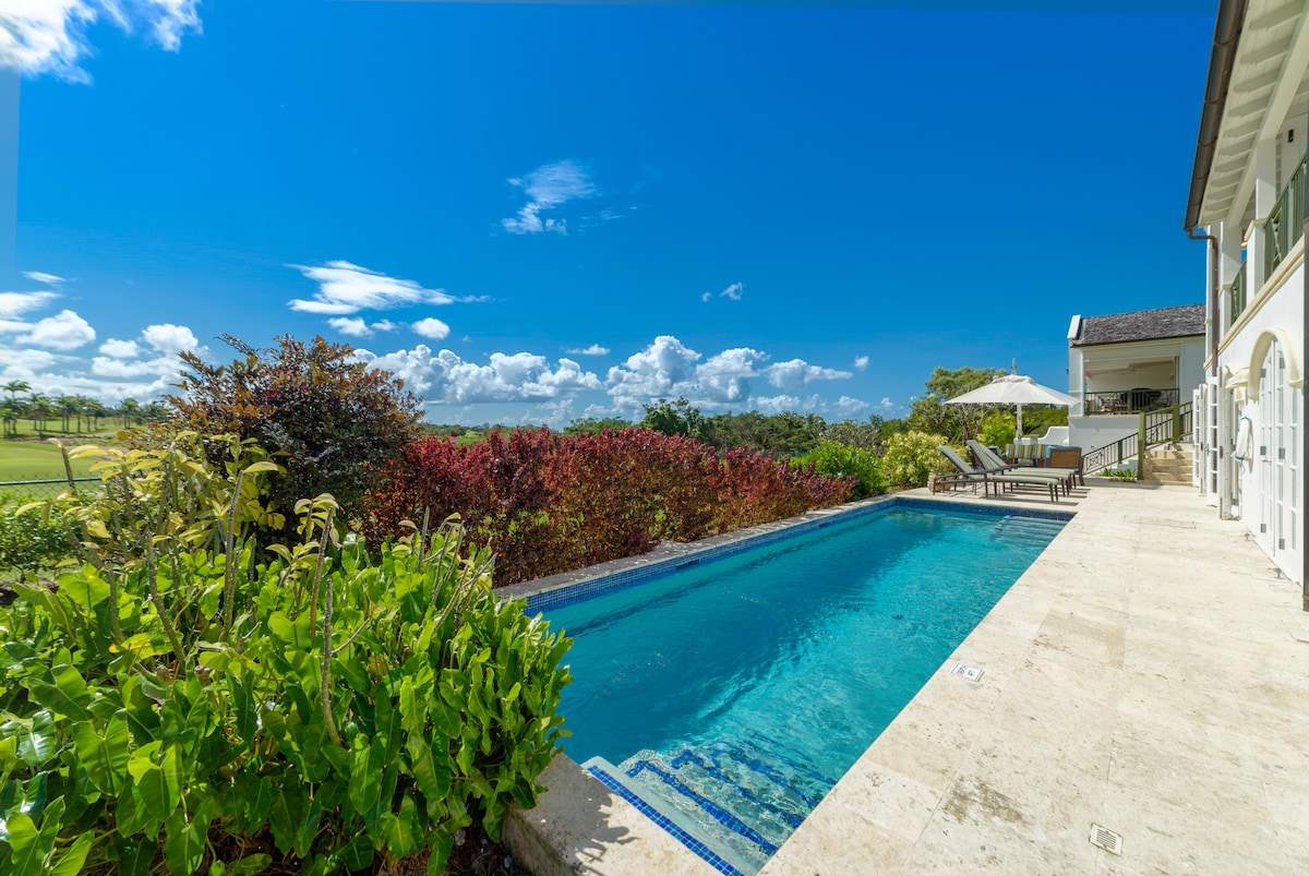 The outdoor space features a private swimming pool surrounded by lush greenery and colorful shrubs. Comfortable lounge chairs are positioned nearby, offering views of the golf course and the coastal landscape under a clear blue sky.
