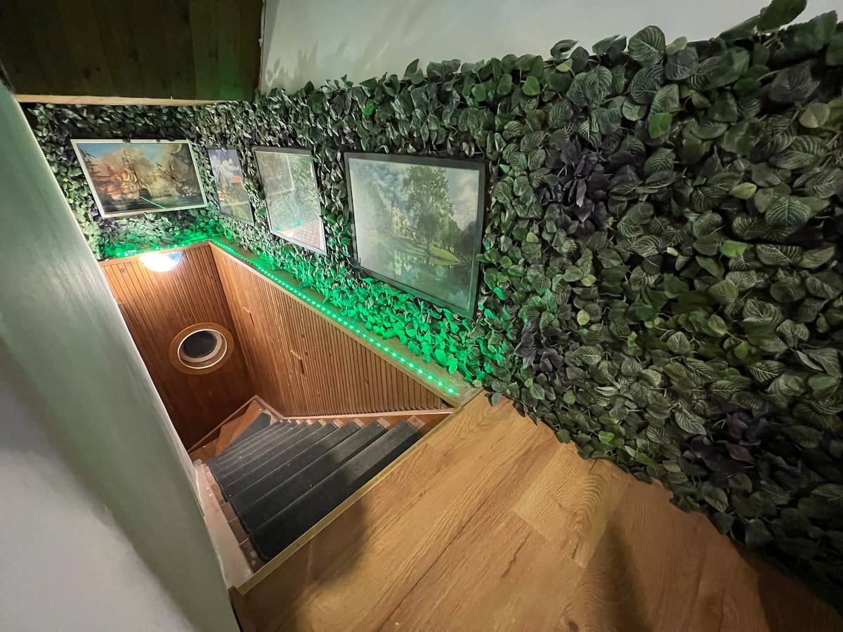 A staircase is adorned with greenery along the walls, creating an inviting ambiance. Framed artwork is visible, complementing the natural decor. Soft lighting enhances the warm tones of the wooden steps, leading down into a welcoming space with a circular window at the base.