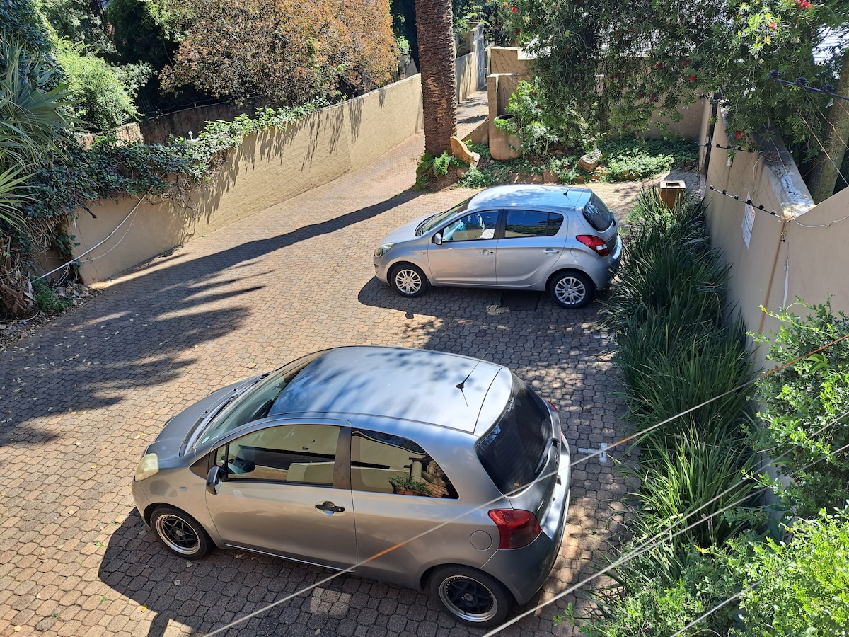A spacious parking area is depicted, accommodating two vehicles. The surrounding greenery and landscaped areas add a natural touch. The driveway features a textured surface, while the background highlights well-maintained walls lined with plants, creating a serene environment.