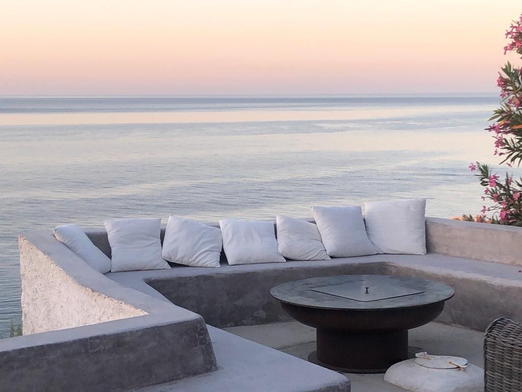 A serene seating area is positioned along a coastal edge, featuring cushioned benches arranged in a curved shape. A stone fire pit is centered within the seating, while the calm sea reflects soft pastel colors of the evening sky.