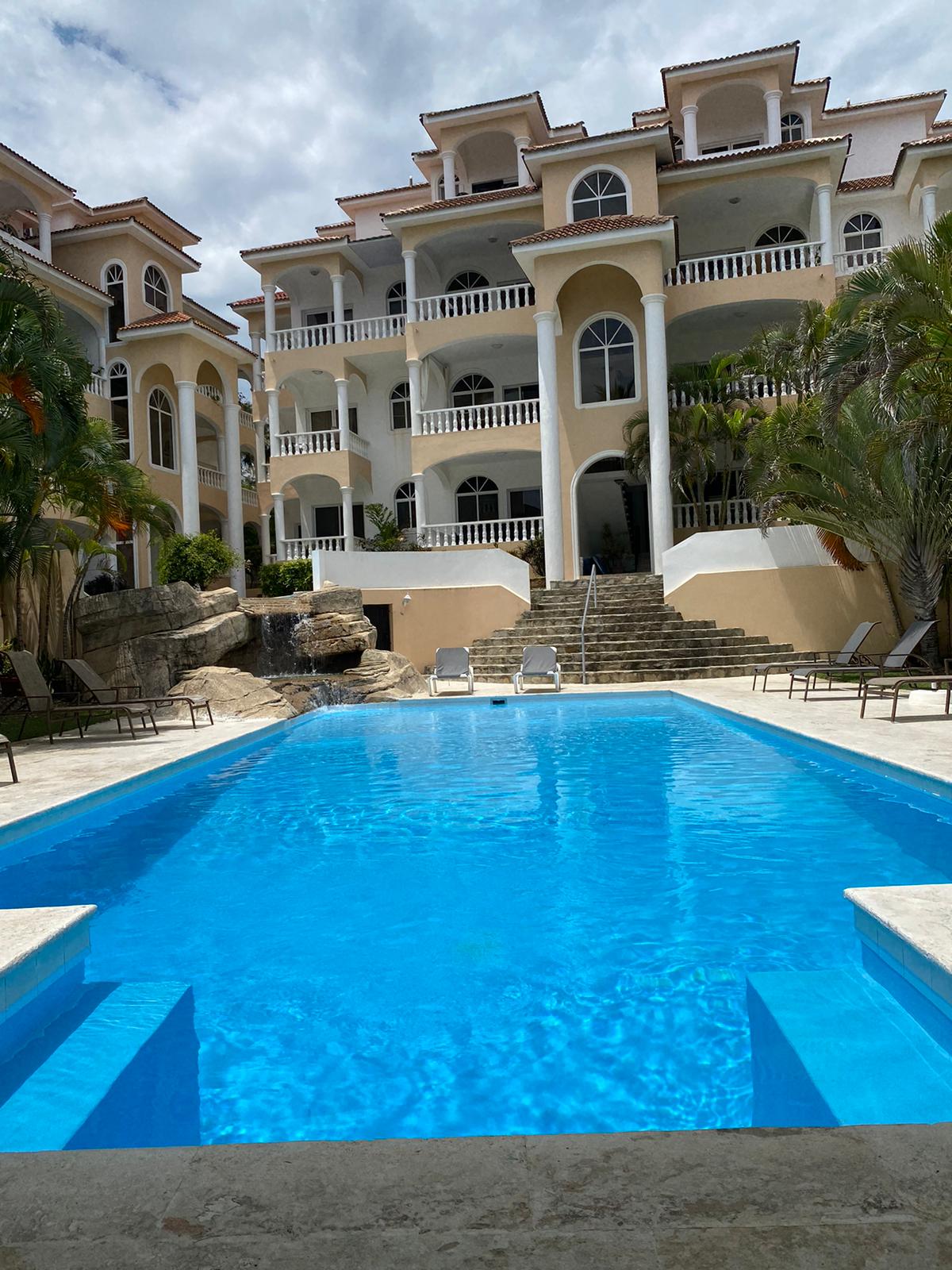 A well-maintained pool area is depicted, featuring clear blue water surrounded by stone and concrete. Comfortable lounge chairs are positioned near the pool's edge. The backdrop showcases a multi-level condominium structure with archways and balconies, framed by lush greenery.