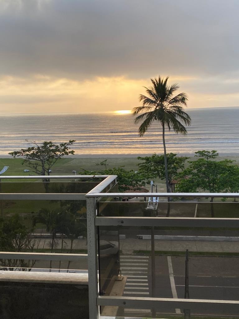 A view of the serene beachfront is captured from a balcony, featuring gentle waves lapping at the shore. A palm tree stands prominently nearby, while the sun rises softly in the background, casting warm hues over the scene.