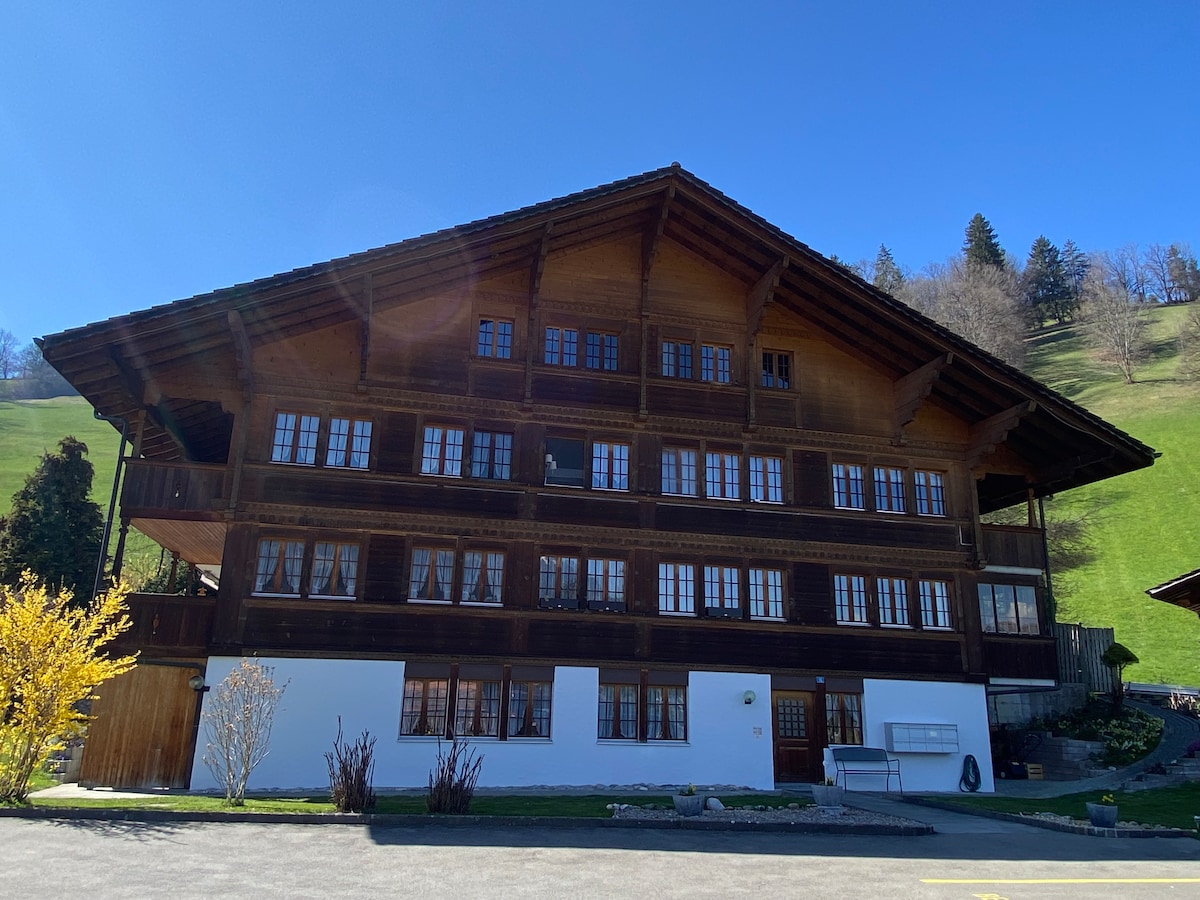 A traditional wooden building is presented with multiple windows, showcasing a blend of dark timber and light-colored lower sections. The well-maintained exterior is surrounded by greenery, with rolling hills rising in the background under a clear blue sky.