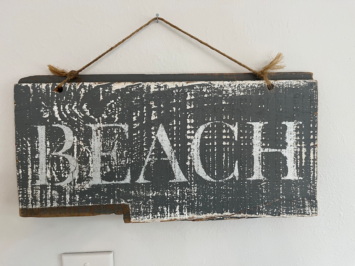 A rustic wall sign hangs from a twine loop, featuring the word 'BEACH' in large, bold white letters against a weathered gray background. The texture of the wood adds to its coastal charm.