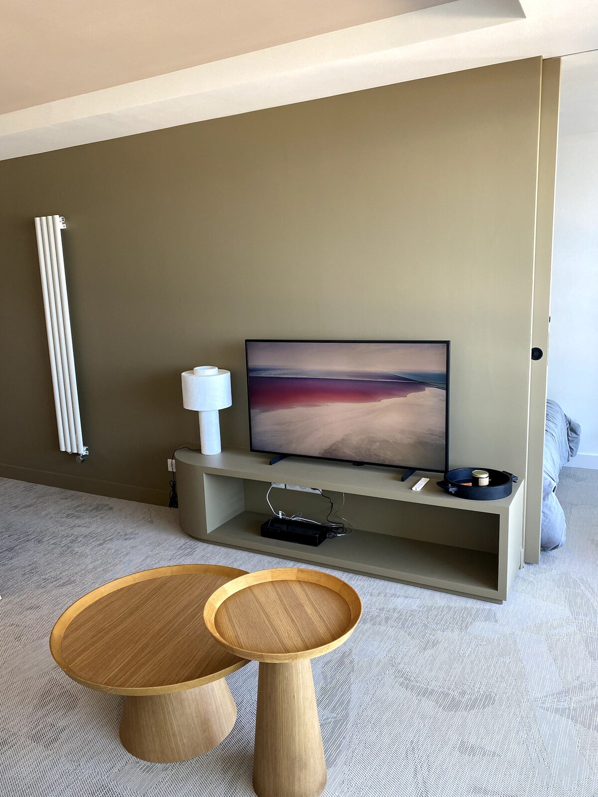 A living area is presented featuring a TV mounted on a simple entertainment unit, complemented by a lamp and small decorative items. Two wooden side tables, one taller than the other, are positioned in front, while a muted green wall adds a calming touch.