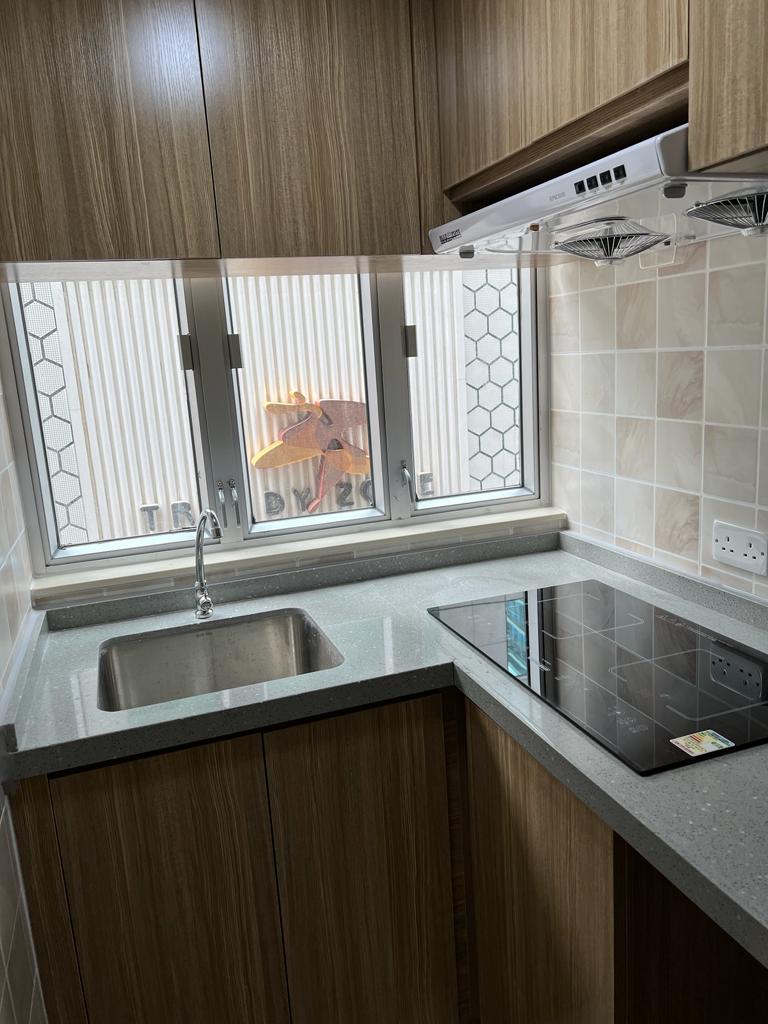 A compact kitchen features wooden cabinetry and a light-colored countertop. A stainless steel sink is positioned below a window, allowing natural light to enter. A glass stovetop is visible beside the sink, with minimalist white tiles enhancing the overall design.