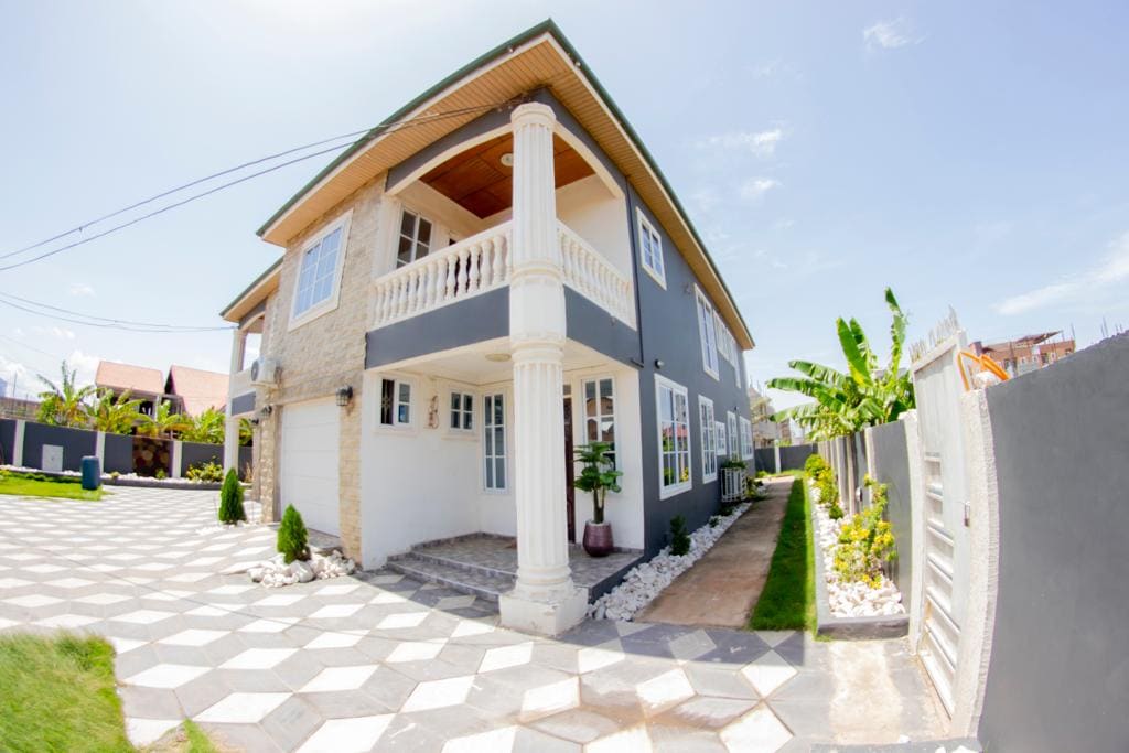 A newly constructed retreat features a two-story structure with a modern design, showcasing large windows and airy balconies. The exterior is complemented by a neatly landscaped yard, with patterned stone pathways and greenery along the sides, creating an inviting entrance.