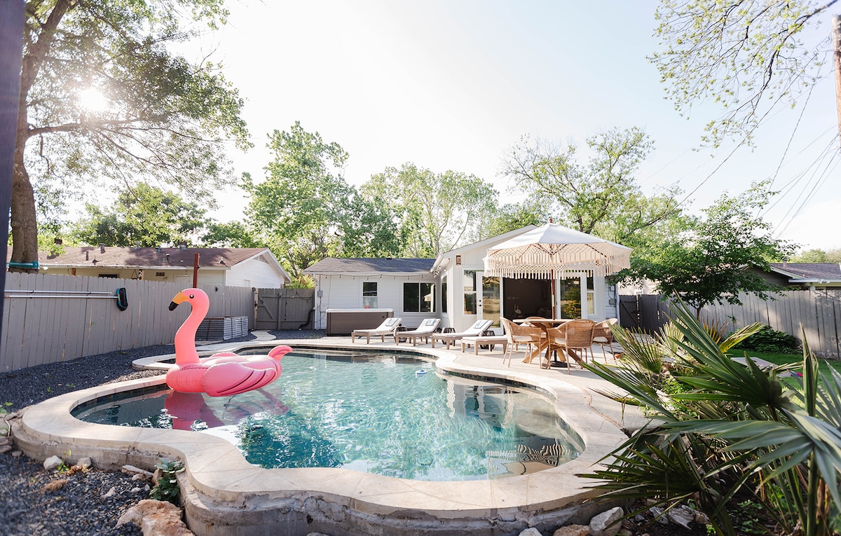 A tropical oasis features a sparkling pool with a pink flamingo float, surrounded by a stone deck and lush greenery. Sun loungers are positioned beside the pool, offering a relaxing space. A shaded outdoor dining area can be seen, inviting leisurely meals al fresco.