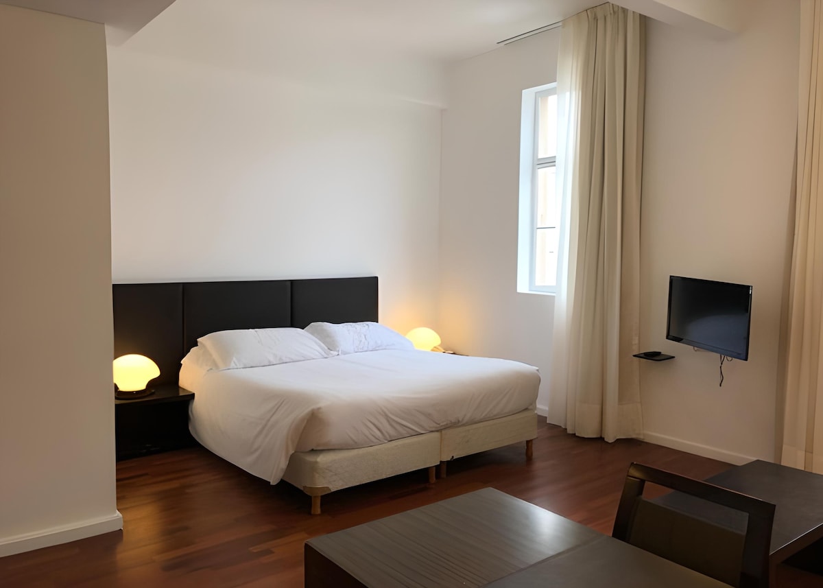 A spacious bedroom features a comfortable bed with soft bedding and two bedside lamps providing gentle illumination. A wall-mounted television is visible across from the bed. Large windows allow natural light to fill the room, and light-colored curtains frame the window.