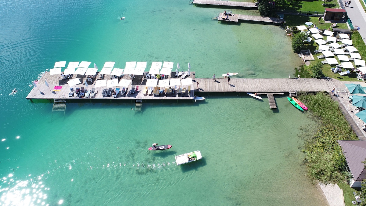 An aerial view of a wooden jetty extending into clear turquoise waters. Sun loungers and umbrellas line the jetty, with people enjoying the water. Kayaks and paddle boards are visible near the jetty, contributing to the serene lakeside atmosphere.
