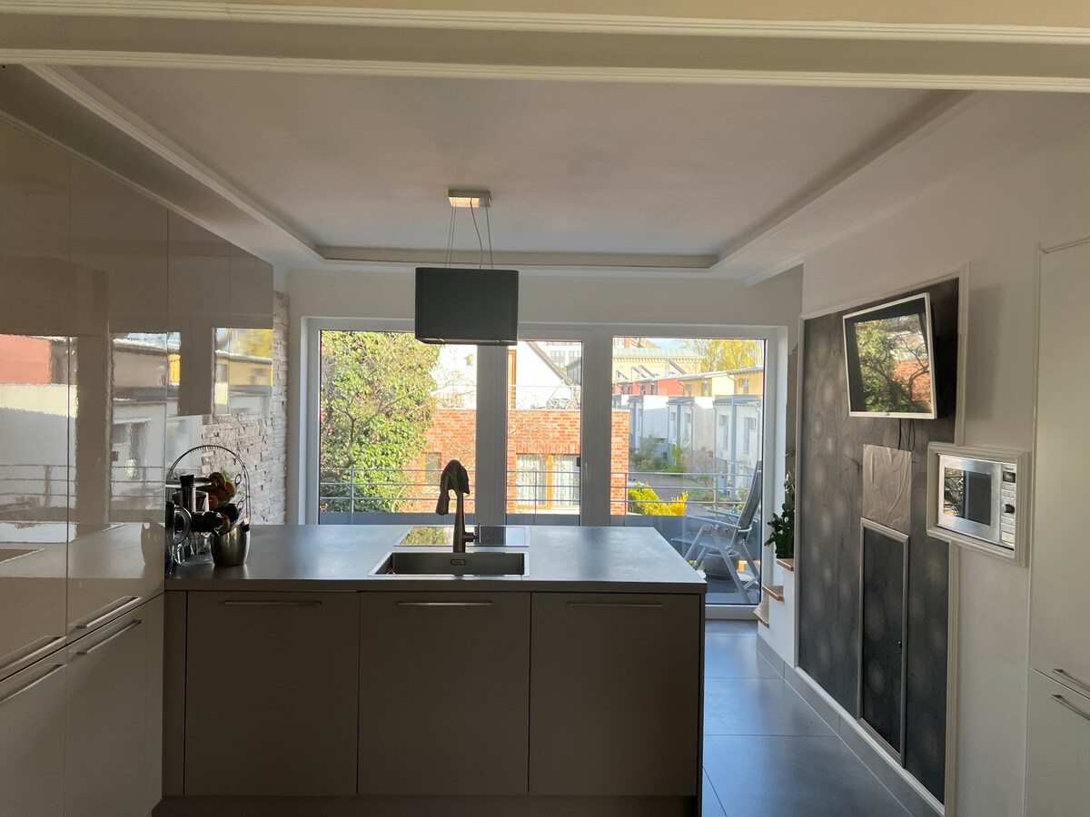 A modern kitchen is presented, featuring a central island with a sink and sleek cabinetry. Large windows offer a view of the outdoor area, allowing natural light to fill the space. A small dining area is also visible, enhancing the functional layout.