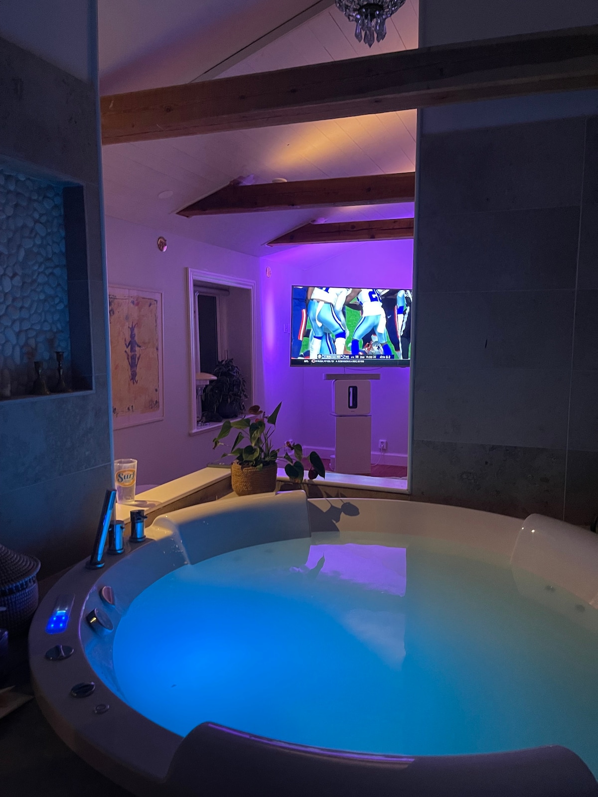 A spacious bathroom features a jacuzzi tub with soft blue lighting, creating a relaxing ambiance. A television is mounted on the wall, displaying a sports event. Natural light is provided by a window, with decorative elements enhancing the modern space.
