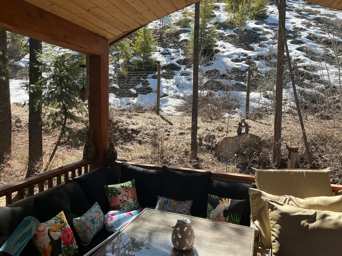 An outdoor enclosed deck features comfortable seating with decorative cushions. A glass table sits at the center, surrounded by trees and a snowy hillside. Natural light filters through the overhead structure, creating a serene space for relaxation.