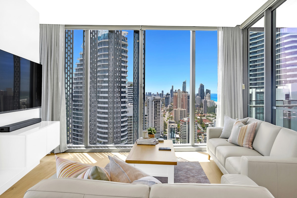 A bright living area showcases large glass windows providing a panoramic view of the city and ocean. Light-colored furnishings include a cozy sofa and a wooden coffee table. Soft curtains frame the windows, allowing natural light to fill the space.
